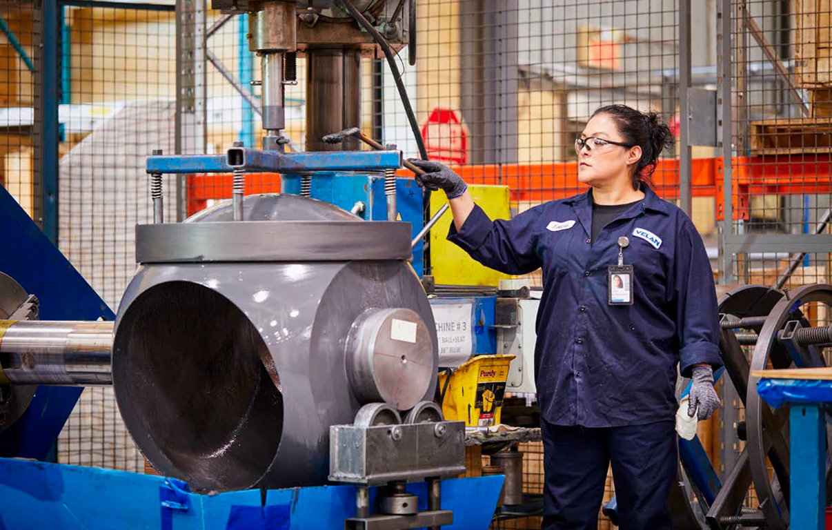 A woman in a navy blue work uniform operates a large industrial machine in a factory. She wears safety glasses, gloves, and has an ID badge. The machine appears to be handling a large metal component. Safety barriers and equipment are visible in the background.
