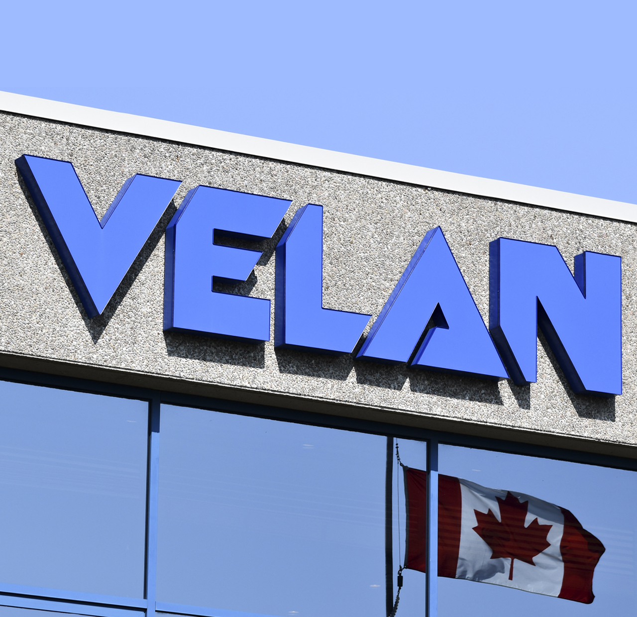L'image montre l'extérieur d'un bâtiment avec le logo « VELAN » en grandes lettres bleues fixé au mur. Sous le logo, un drapeau canadien est visible, suggérant que l'entreprise pourrait avoir un lien avec le Canada. Le ciel est clair et bleu en arrière-plan.